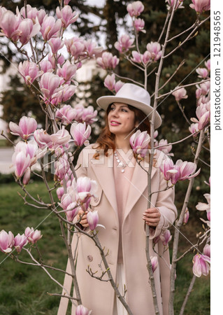 Woman magnolia flowers, surrounded by blossoming trees., hair down, white hat, wearing a light coat. Captured during spring, showcasing natural beauty and seasonal change. Woman magnolia flowers, surrounded by blossoming trees., hair down, white hat, wearing a light coat. Captured during spring, showcasing natural beauty and seasonal change. 121948565