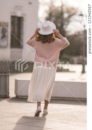 A woman wearing a white hat and a pink sweater stands in front of a building 121948567