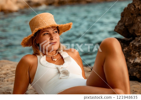 Woman Beach Sunbathing Summer - A woman in a white swimsuit relaxes on a rocky beach while wearing a straw hat, enjoying the summer sun. Woman Beach Sunbathing Summer - A woman in a white swimsuit relaxes on a rocky beach while wearing a straw hat, enjoying the summer sun. 121948635