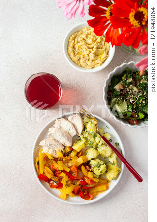 A balanced meal of boiled potatoes with baked vegetables and chicken breast, paired with a fresh tomato and cucumber salad topped with seeds, and a refreshing raspberry compote 121948864