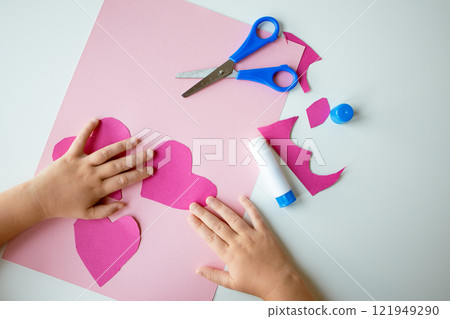 Childs hands making pink paper hearts with scissors and glue on white table Childs hands making pink paper hearts with scissors and glue on white table 121949290