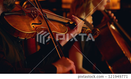 A close-up of musicians playing string instruments in an orchestra, focusing on a violinist and a cellist. The warm lighting highlights the wood of the instruments A close-up of musicians playing string instruments in an orchestra, focusing on a violinist and a cellist. The warm lighting highlights the wood of the instruments 121949771