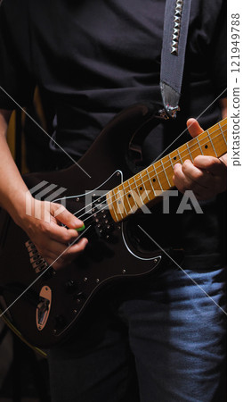 Musician in black clothes and blue jeans playing a black electric guitar with maple deck, green plectrum visible, studded guitar strap attached 121949788