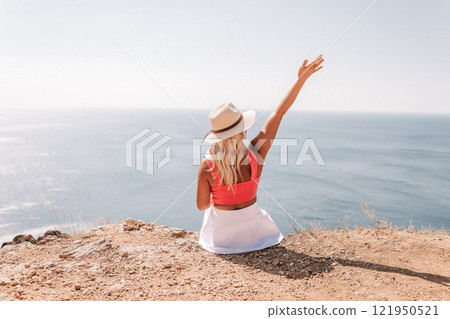 A woman in a pink top and white skirt is sitting on a beach, with her arms raised in the air. Concept of relaxation and enjoyment of the beautiful surroundings. A woman in a pink top and white skirt is sitting on a beach, with her arms raised in the air. Concept of relaxation and enjoyment of the beautiful surroundings. 121950521