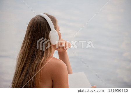 A woman wearing headphones is looking out at the water. The scene is calm and peaceful, with the woman seemingly lost in thought. A woman wearing headphones is looking out at the water. The scene is calm and peaceful, with the woman seemingly lost in thought. 121950580