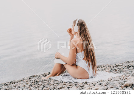 A woman is sitting on the beach with a laptop and headphones on. She is wearing a white bikini and she is enjoying her time by the water. 121950581