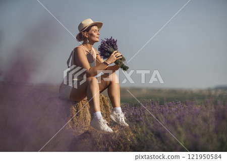woman sitting in a field of lavender and wearing a straw hat. She is smiling and holding a bouquet of flowers. Scene is peaceful and serene, as the woman is surrounded by the beauty of nature 121950584