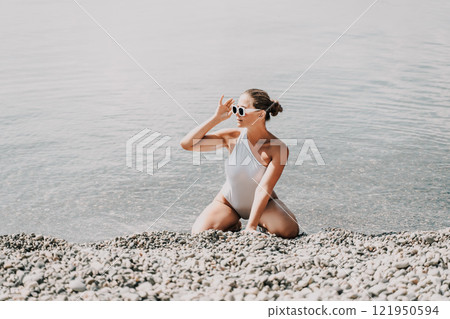 A woman in a white bikini is sitting on the beach, looking out at the water. She is wearing sunglasses and she is enjoying the view. 121950594