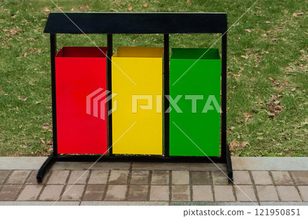 Outdoor garbage bin with three containers for separate collection, painted red, yellow and green, installed on pedestrian path in public park, on green lawn background. Black metal frame, with a roof Outdoor garbage bin with three containers for separate collection, painted red, yellow and green, installed on pedestrian path in public park, on green lawn background. Black metal frame, with a roof 121950851