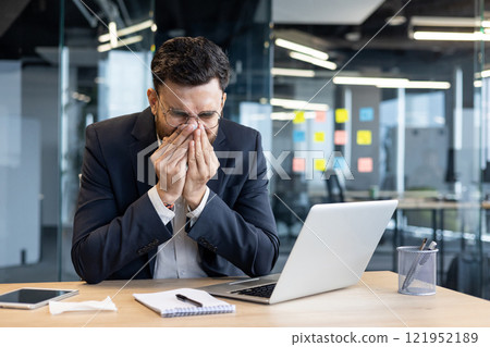Sick man at workplace, has runny nose sneezing, allergic attack. Businessman inside office working with laptop. 121952189