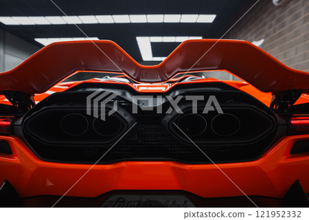 The image captures the rear of a Lamborghini Revuelto indoors, featuring dual exhausts, vibrant orange color, angular spoiler, and sleek aerodynamic lines. The image captures the rear of a Lamborghini Revuelto indoors, featuring dual exhausts, vibrant orange color, angular spoiler, and sleek aerodynamic lines. 121952332