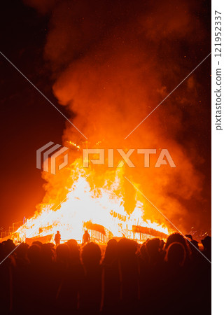 A towering bonfire illuminates the night sky, surrounded by silhouetted spectators. Sparks fly as the flames cast an orange glow in the Black Rock Desert. 121952337