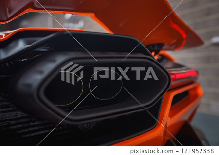 The image captures the vibrant orange rear exhaust of a Lamborghini Revuelto, highlighting its angular design and dual exhaust pipes against a blurred brick wall. 121952338