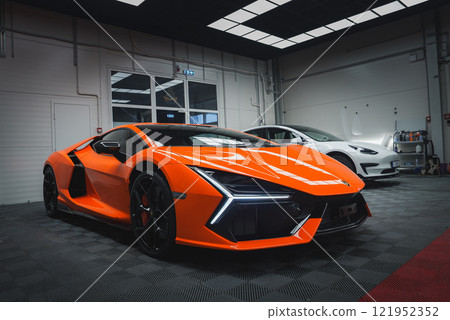 An orange Lamborghini Revuelto is parked indoors, showcasing its sleek design and LED headlights. A checkered floor and grid ceiling enhance the setting. 121952352