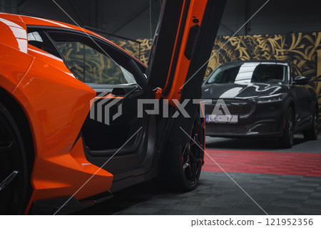 An orange Lamborghini Revuelto with its scissor door open is displayed indoors on a checkered floor, set against gold patterned wallpaper. A gray Ford Mustang Mach E is visible in the background. An orange Lamborghini Revuelto with its scissor door open is displayed indoors on a checkered floor, set against gold patterned wallpaper. A gray Ford Mustang Mach E is visible in the background. 121952356