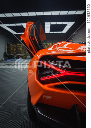 The image captures the rear section of an orange Lamborghini Revuelto indoors, highlighting its aerodynamic design, distinctive taillights, and open scissor door. 121952360