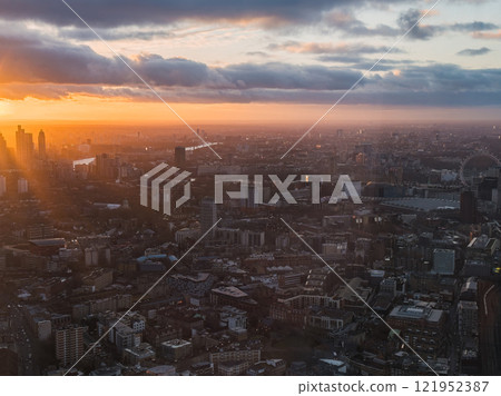 The image shows an aerial view of London during sunset, featuring the London Eye and River Thames. The warm glow highlights the urban landscape and clouds. The image shows an aerial view of London during sunset, featuring the London Eye and River Thames. The warm glow highlights the urban landscape and clouds. 121952387