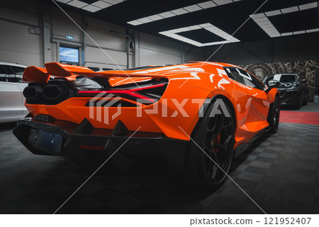 An orange Lamborghini Revuelto is displayed in a well lit showroom, featuring sharp lines, distinctive rear lights, and dual exhausts on checkered flooring. 121952407