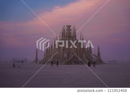 A large wooden dome with spires and latticework stands in the desert. The sky is pink and purple, and participants walk around the installation. A large wooden dome with spires and latticework stands in the desert. The sky is pink and purple, and participants walk around the installation. 121952408