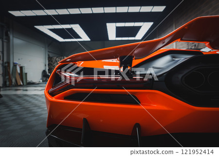 The image captures the rear section of a Lamborghini Revuelto, focusing on its taillights and rear diffuser. The vibrant orange car is in a modern showroom. 121952414