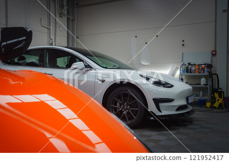 A vibrant orange Lamborghini Revuelto is partially visible in the foreground, while a white Tesla Model 3 is parked in a garage, surrounded by car care products. 121952417