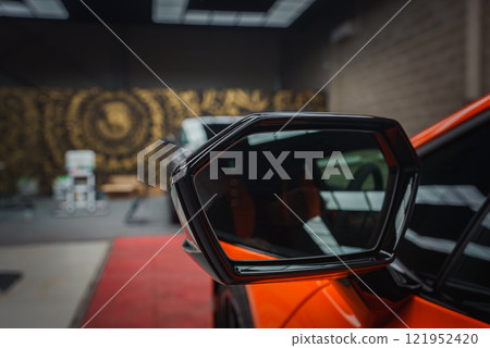 A vibrant orange Lamborghini Revuelto's side mirror is showcased against a dark, industrial style background with intricate gold patterns. A vibrant orange Lamborghini Revuelto's side mirror is showcased against a dark, industrial style background with intricate gold patterns. 121952420