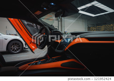 The image features the sleek interior of a Lamborghini Revuelto, highlighting vibrant orange accents against a dark backdrop. An open scissor door reveals part of the exterior and another car in a 121952424