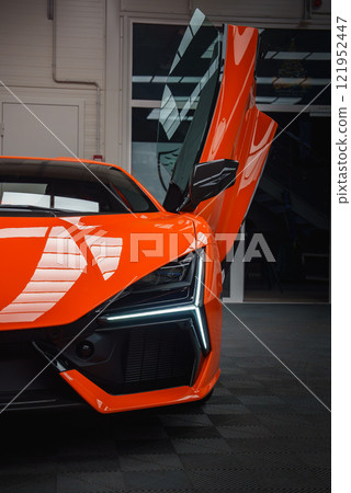 The image features the front section of an orange Lamborghini Revuelto indoors, highlighting its angular design, LED headlights, and open scissor door. The image features the front section of an orange Lamborghini Revuelto indoors, highlighting its angular design, LED headlights, and open scissor door. 121952447