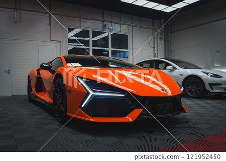 An orange Lamborghini Revuelto is parked indoors on a checkered floor, showcasing its angular headlights and aerodynamic body. A white car is beside it. 121952450