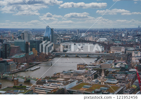The image captures London's urban landscape with the River Thames, the Shard, and the London Eye. Modern and historic architecture blend under a cloudy sky. The image captures London's urban landscape with the River Thames, the Shard, and the London Eye. Modern and historic architecture blend under a cloudy sky. 121952456