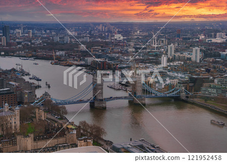 View of Tower Bridge spanning the River Thames in London at sunset. The sky is painted in orange and red hues, reflecting on the river below. 121952458