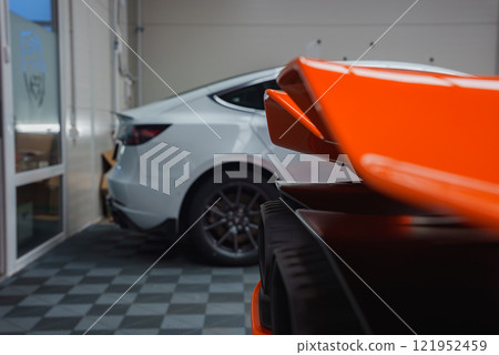 The image shows a close up of a Lamborghini Revuelto's vibrant orange rear wing in an indoor showroom, with tiled flooring and a white car nearby. 121952459
