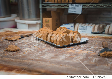 A wooden table dusted with flour holds a tray of sliced brown confectionery. Shelves in the background display containers and a sign reading 'Vanilla and Raisin'. 121952460