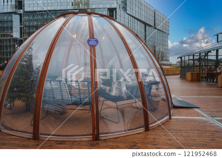 A transparent igloo style dome on a wooden deck in London features cozy seating, blankets, and a small Christmas tree, with a modern glass building in the background. A transparent igloo style dome on a wooden deck in London features cozy seating, blankets, and a small Christmas tree, with a modern glass building in the background. 121952468