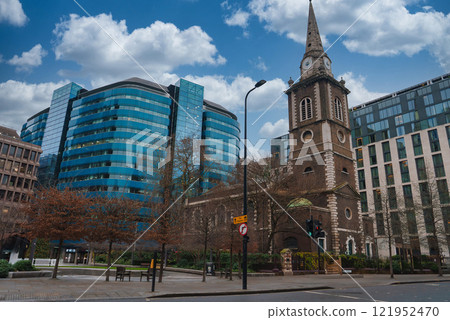 A traditional church with a tall spire and clock stands amidst modern glass buildings in London. Bare trees line the street under a clear sky with clouds. 121952470