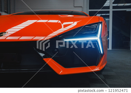 The image captures the front end of a Lamborghini Revuelto indoors, focusing on its angular design, vibrant orange color, and distinctive LED headlight. 121952471