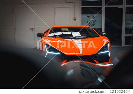 A vibrant orange Lamborghini Revuelto is showcased indoors, highlighting its angular design, LED headlights, and emblem. Glass doors and walls surround the car. 121952494