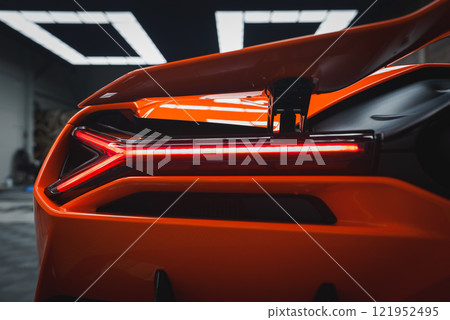 The image captures the rear section of a Lamborghini Revuelto, focusing on its vibrant orange color and illuminated red taillight in an indoor environment. 121952495