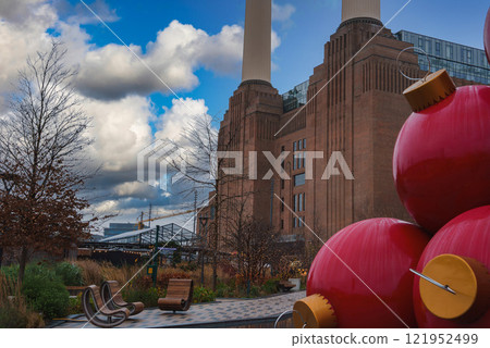 Battersea Power Station's chimneys rise against a partly cloudy sky, with oversized red Christmas ornaments in the foreground, amid winter landscaping. Battersea Power Station's chimneys rise against a partly cloudy sky, with oversized red Christmas ornaments in the foreground, amid winter landscaping. 121952499