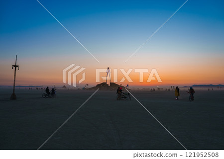 A large wooden art installation stands in a desert at sunset, surrounded by bicyclists. The sky transitions from orange to blue, with mountains in the distance. 121952508
