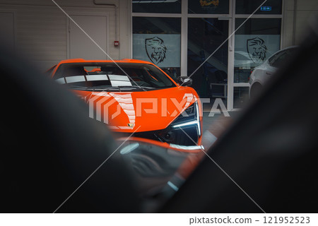 A vibrant orange Lamborghini Revuelto is showcased indoors, featuring LED headlights and emblem. Glass doors with lion logos are visible in the background. 121952523