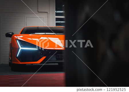A close up view of a Lamborghini Revuelto in vibrant orange, highlighting its angular front, distinctive LED headlights, and sleek lines indoors. 121952525