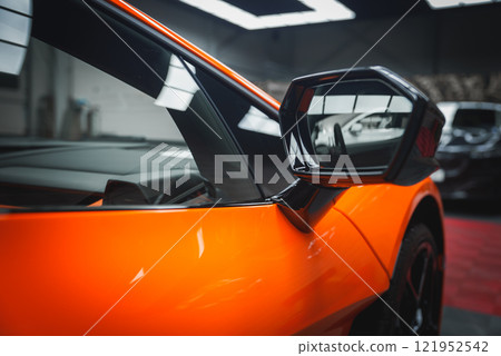 A close up view of a Lamborghini Revuelto's angular side mirror and vibrant orange bodywork, set in an indoor showroom with a modern ceiling design. 121952542