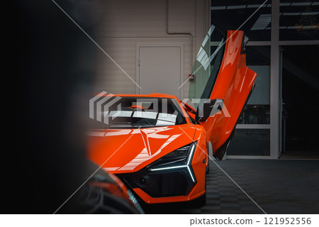 Close up of an orange Lamborghini Revuelto indoors, showcasing its open scissor door, aggressive headlights, and sleek design against a modern backdrop. 121952556