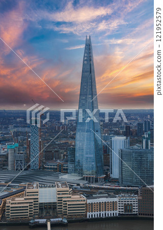 The Shard skyscraper stands tall against a vibrant sunset sky in London. Modern buildings and cranes surround it, with the River Thames in the foreground. 121952579