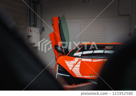A vibrant orange Lamborghini Revuelto is showcased indoors with its scissor door open. The setting includes a brick wall and electrical panels in the background. 121952580