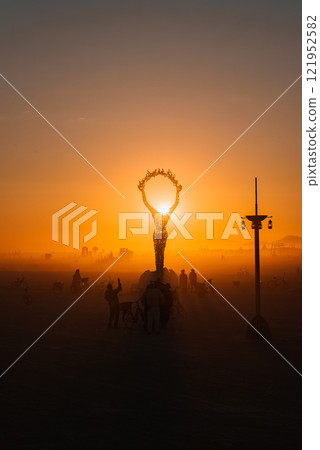 A large human figure sculpture with raised arms is silhouetted against a sunset at a desert festival, surrounded by participants and bicycles. A large human figure sculpture with raised arms is silhouetted against a sunset at a desert festival, surrounded by participants and bicycles. 121952582