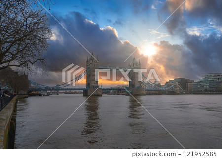 Tower Bridge stands over the River Thames in London, with the sun breaking through clouds. Bare trees and modern buildings frame the winter scene. Tower Bridge stands over the River Thames in London, with the sun breaking through clouds. Bare trees and modern buildings frame the winter scene. 121952585