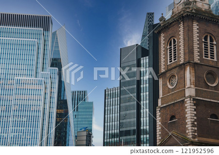 A traditional brick church with arched windows and a clock face stands beside sleek glass skyscrapers, under a clear blue sky in London during Christmas. A traditional brick church with arched windows and a clock face stands beside sleek glass skyscrapers, under a clear blue sky in London during Christmas. 121952596