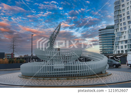 A modern spiral sculpture of white metal slats stands against contemporary buildings in London, with a vibrant sunset sky enhancing the urban scene. A modern spiral sculpture of white metal slats stands against contemporary buildings in London, with a vibrant sunset sky enhancing the urban scene. 121952607
